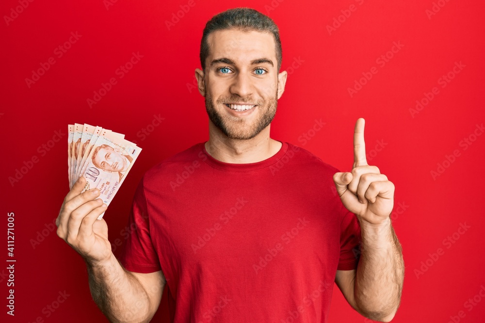 Young caucasian man holding 10 singapore dollars banknotes smiling with an idea or question pointing finger with happy face, number one Young caucasian man holding 10 singapore dollars banknotes smiling with an idea or question pointing finger with happy face, number one