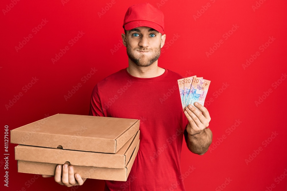 Young caucasian man holding delivery box and swiss franc banknotes puffing cheeks with funny face. mouth inflated with air, catching air. Young caucasian man holding delivery box and swiss franc banknotes puffing cheeks with funny face. mouth inflated with air, catching air.