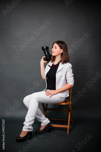 Attractive girl photographer sits on a chair with a camera in her hands in the studio on a gray background.