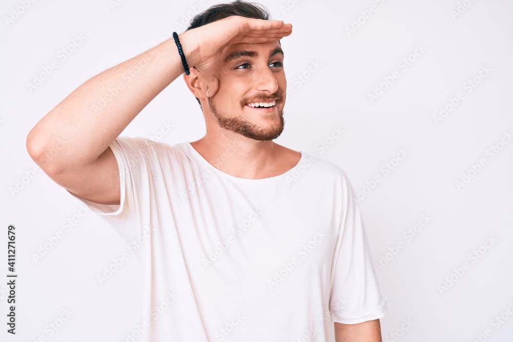 Young handsome man wearing casual white tshirt very happy and smiling looking far away with hand over head. searching concept. Young handsome man wearing casual white tshirt very happy and smiling looking far away with hand over head. searching concept.