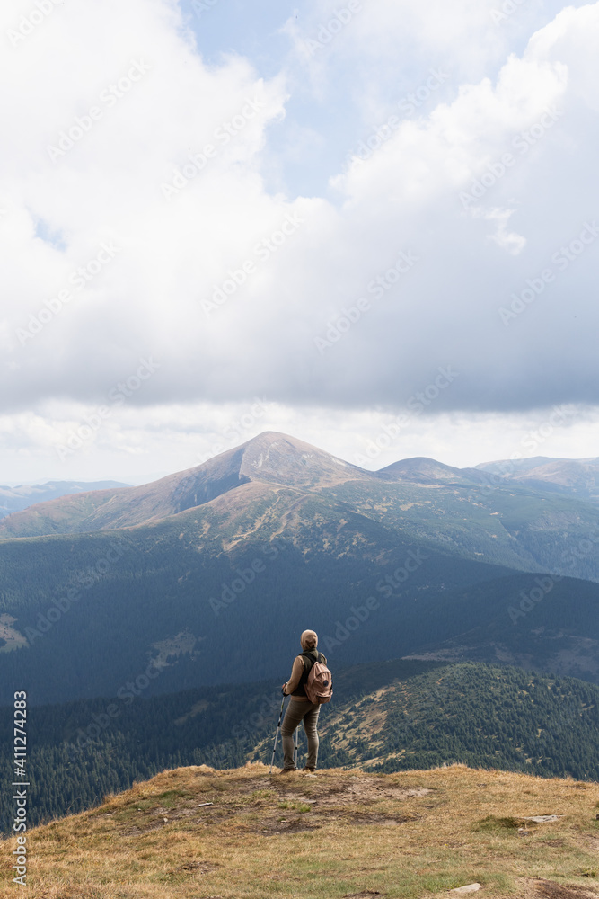 Woman hiking with backpack and trekking poles. Nature tourism in Ukrainian Carpathian mountains. Hoverla mountain peak in the background.