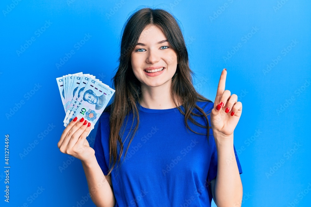 Young beautiful caucasian girl holding 10 yuan chinese banknotes smiling with an idea or question pointing finger with happy face, number one Young beautiful caucasian girl holding 10 yuan chinese banknotes smiling with an idea or question pointing finger with happy face, number one