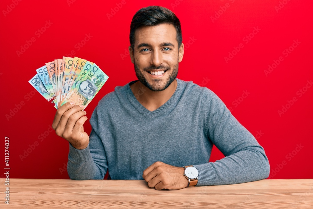 Handsome hispanic man holding australian dollars looking positive and happy standing and smiling with a confident smile showing teeth Handsome hispanic man holding australian dollars looking positive and happy standing and smiling with a confident smile showing teeth