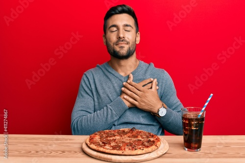 Handsome hispanic man eating tasty pepperoni pizza smiling with hands on chest, eyes closed with grateful gesture on face. health concept.