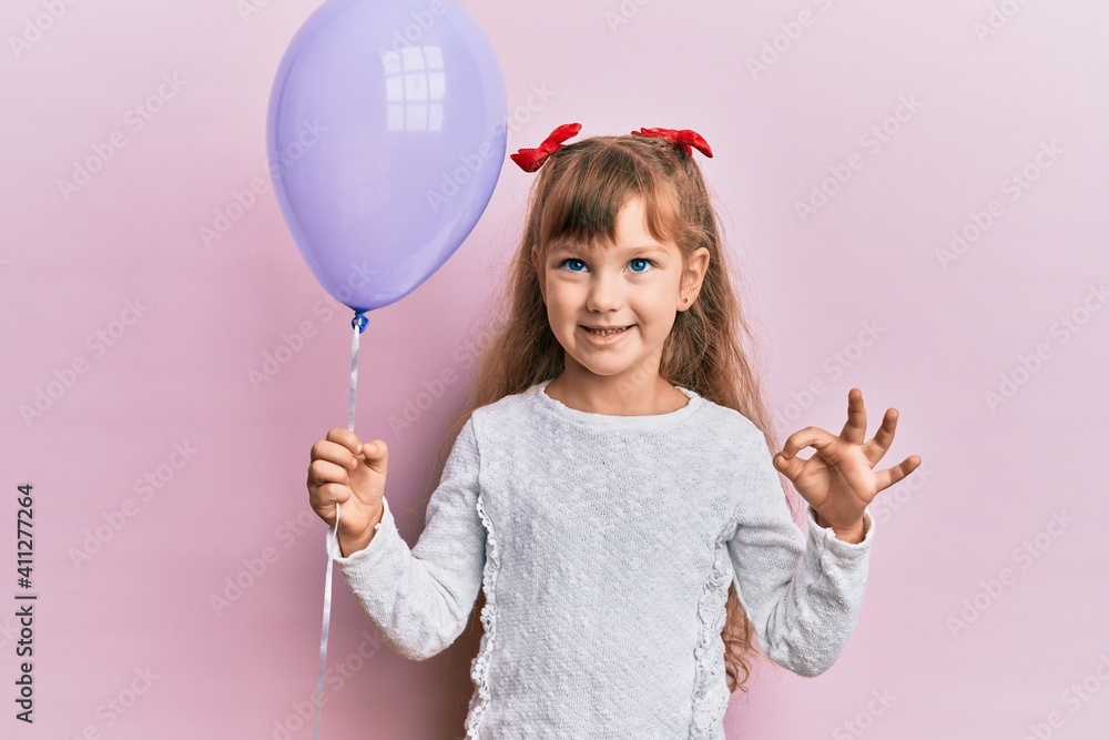 Little caucasian girl kid holding purple balloon doing ok sign with fingers, smiling friendly gesturing excellent symbol