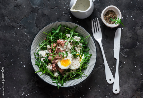 Salad with arugula, canned tuna, boiled egg and yogurt, mustard dressing on a dark background, top view