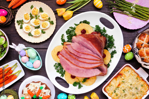 Traditional Easter ham dinner. Above view table scene on a dark wood background. Ham, scalloped potatoes, eggs, hot cross buns, carrot cake and vegetables.