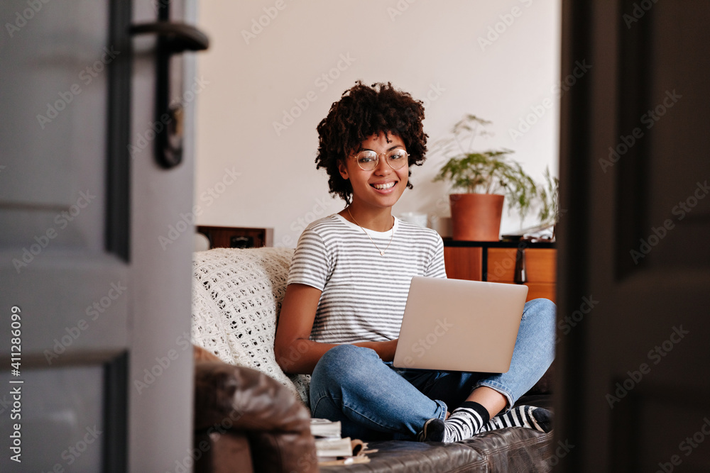 Cute girl in glasses looking at camera. Woman in white t-shirt is resting on sofa and holding laptop Cute girl in glasses looking at camera. Woman in white t-shirt is resting on sofa and holding laptop