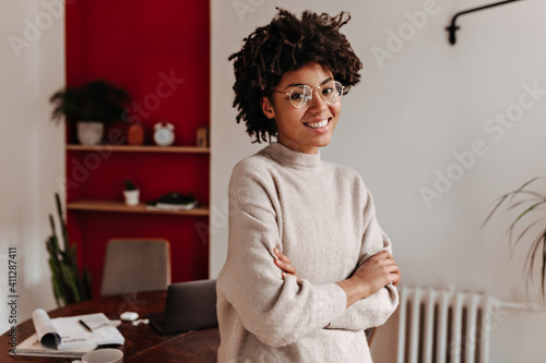 Brown-eyed woman in glasses smiles sweetly and stands at workplace with her arms crossed