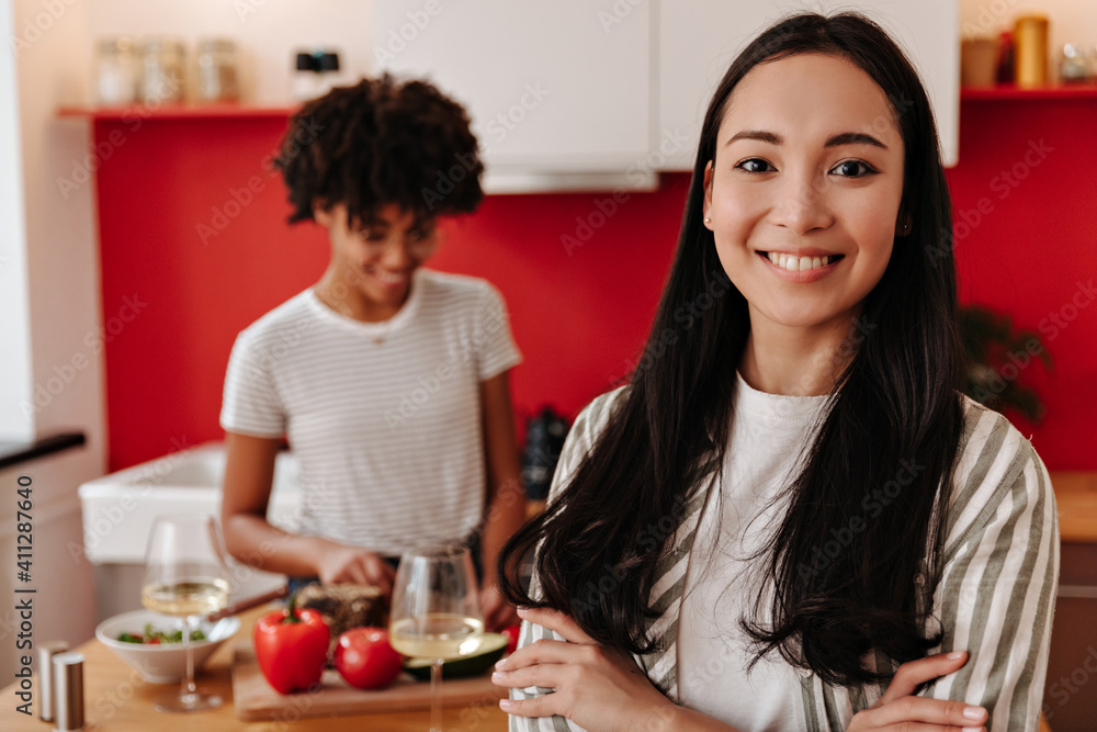 Positive girlfriends are posing in kitchen. Asian looks into camera. Girl in glasses smiles and cuts vegetables