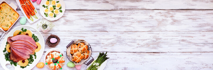 Traditional Easter ham dinner. Overhead view corner border on a white wood banner background with copy space. Ham, scalloped potatoes, vegetables, eggs, hot cross buns and carrot cake.
