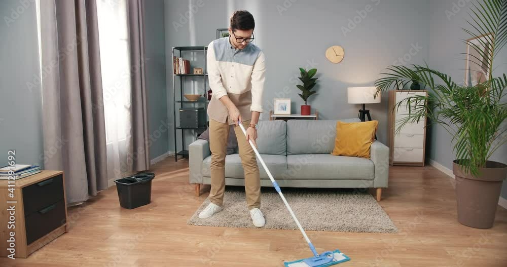 Caucasian handsome young serious male in glasses washing floor using mop in modern living room at home. Man cleaning house alone, housekeeping and householding, clean up, domestic task concept Caucasian handsome young serious male in glasses washing floor using mop in modern living room at home. Man cleaning house alone, housekeeping and householding, clean up, domestic task concept