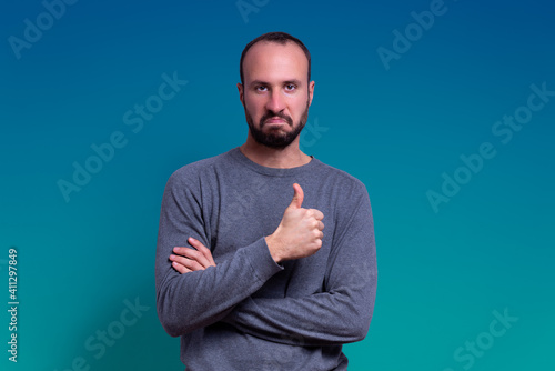 young man with beard and casual clothing giving thumbs up as a gesture of approval with serious and impressed expression on blue fading background. expression of pride and winner