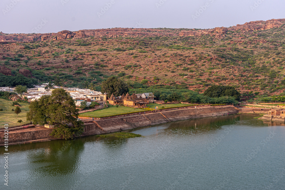 Badami, Karnataka, India - November 7, 2013: Wide area view with Brown stone Hondadkatte Hanuman Temple on NW-shore of Agasthya Lake. Green foliage in back with brown hills. Sunset twilight.