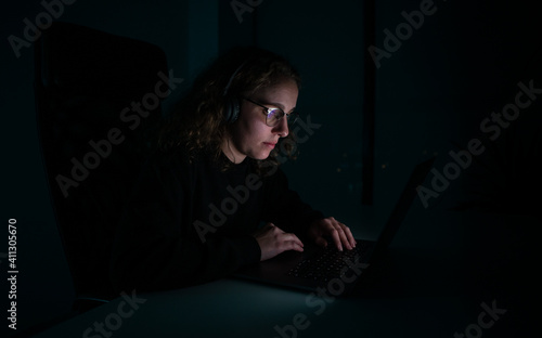 Woman with headphones typing on her laptop in the dark of the night. Overwork.