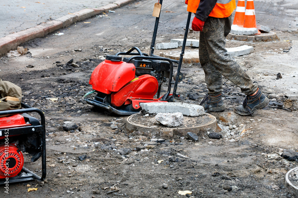 A road worker repairs and installs sewers on the road using a compactor plate and a petrol generator.