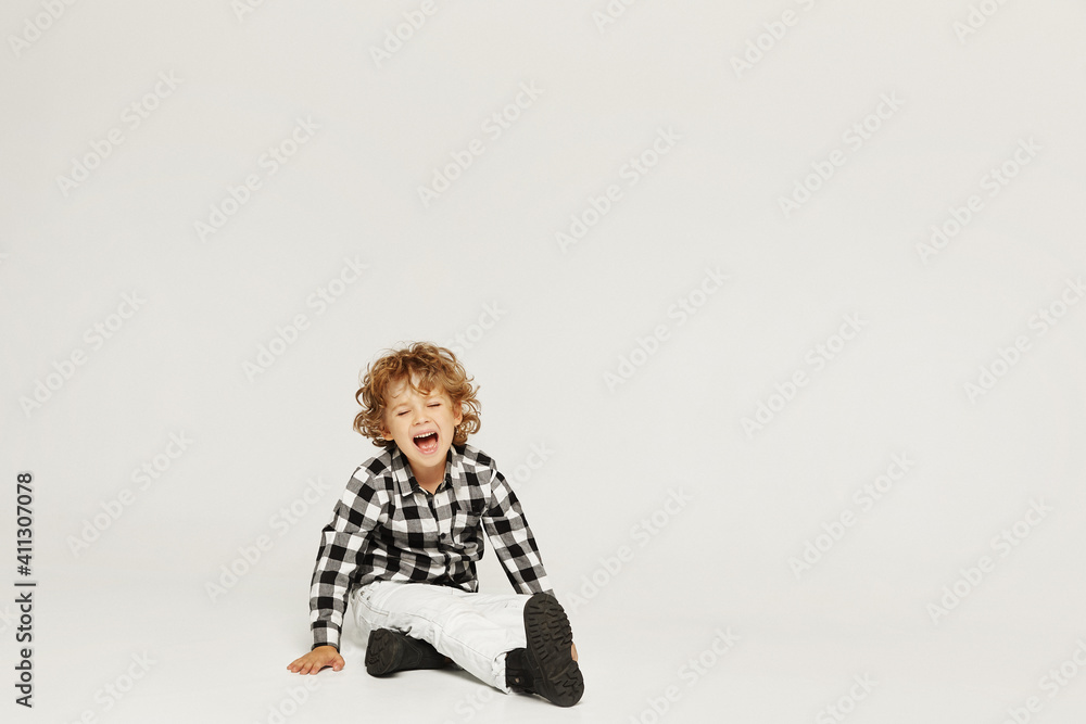 Crying curly little boy sits on the floor and keeping hand on his leg. A small kid broke his leg, isolated over gray background Crying curly little boy sits on the floor and keeping hand on his leg. A small kid broke his leg, isolated over gray background