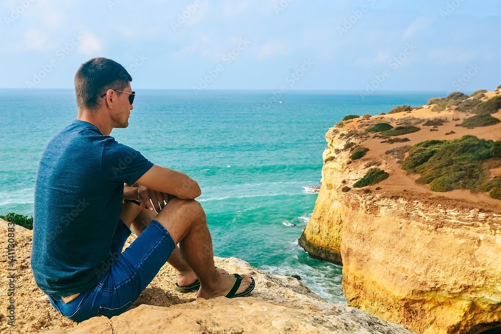 one slender handsome guy with glasses sits on stones on a rock in Portugal. back view of the ocean. one slender handsome guy with glasses sits on stones on a rock in Portugal. back view of the ocean.