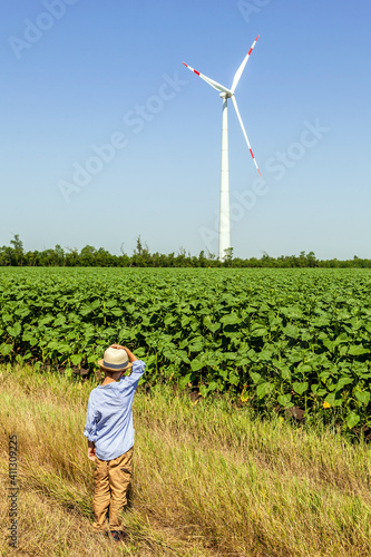 A child from a dirt road looks at wind turbines against a background of cloudy blue sky and green field