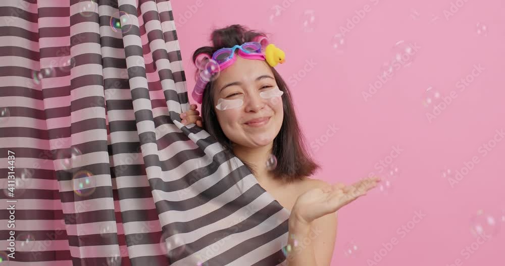 Spa and body washing concept. Happy young Asian woman keeps palm raised catches bubbles has good mood while taking shower isolated over pink background enjoys hygiene procedures at bathroom. Spa and body washing concept. Happy young Asian woman keeps palm raised catches bubbles has good mood while taking shower isolated over pink background enjoys hygiene procedures at bathroom.
