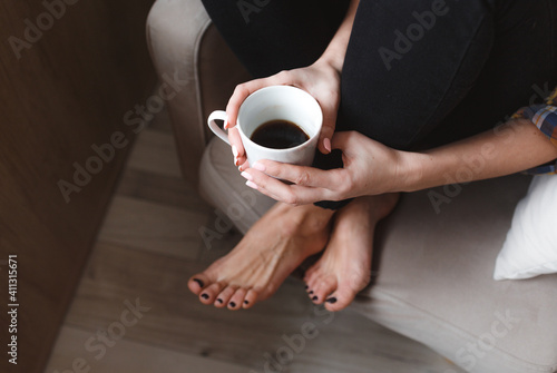 A young woman with a mobile phone is sitting in a comfortable chair by the window of the house and drinking tea. Home interior design. Freelancer