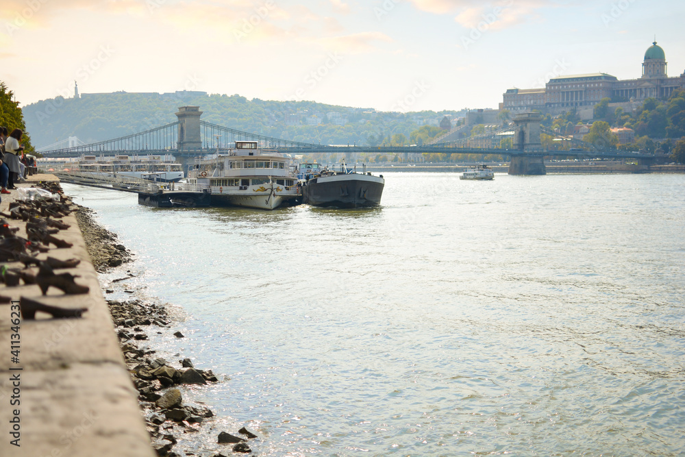 Boats line the banks of the Danube River near the Shoes on the Danube memorial with the Chain Bridge and Royal Palace behind, in Budapest, Hungary