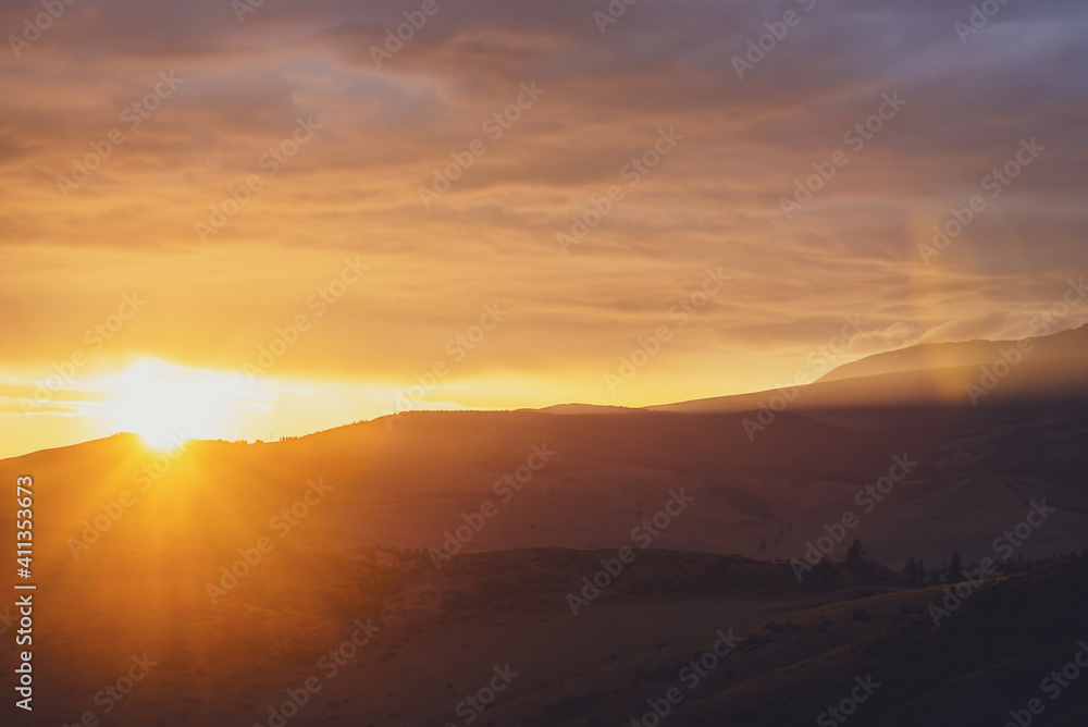 Atmospheric landscape with silhouettes of mountains with trees on background of dawn sky with sun circle and orange sun rays. Colorful nature scenery with sunset or sunrise of illuminating color. Atmospheric landscape with silhouettes of mountains with trees on background of dawn sky with sun circle and orange sun rays. Colorful nature scenery with sunset or sunrise of illuminating color.