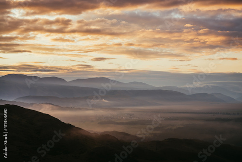 Scenic mountain landscape with golden low clouds above village among mountains silhouettes under dawn cloudy sky. Atmospheric alpine scenery of countryside in low clouds in sundown illuminating color. © Daniil