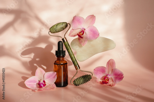 Photo for the spa salon. Roller and Guasha scraper for facial massage  massage oil  black hot spa stones  pink orchids  white towel on a pink background.  Balance and levitation trending style
