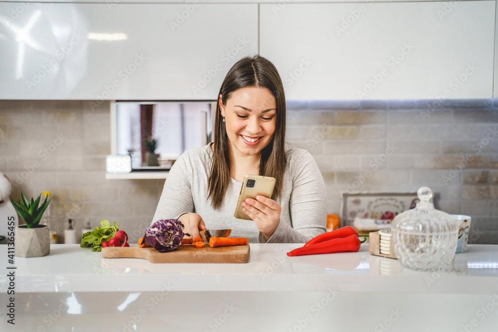 Portrait of young adult caucasian woman in the kitchen using mobile phone browsing internet checking recipe online while cutting vegetable preparing meal - copy space real people modern healthy living Portrait of young adult caucasian woman in the kitchen using mobile phone browsing internet checking recipe online while cutting vegetable preparing meal - copy space real people modern healthy living