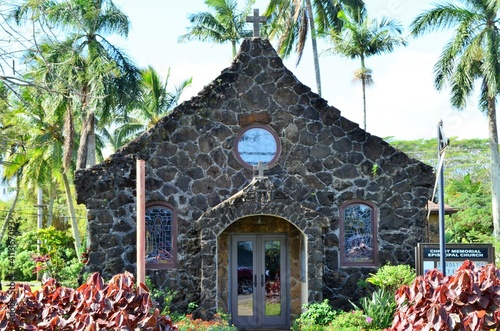 A stone front church sits amongst large swaying palm trees and beautiful tropical flowers in the wonderful island of Hawaii
