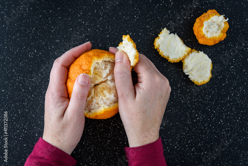 Woman’s hands peeling a fresh large orange with wrinkled and textured skin, black cutting board
