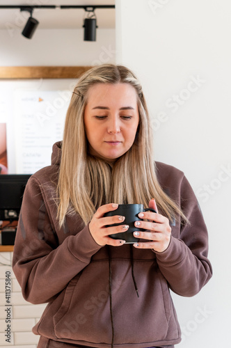 Young girl in a brown tracksuit stands in a cafe and holds hot designer tea in her hands.