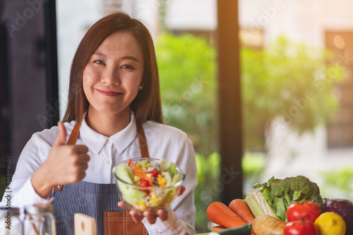 Closeup image of a beautiful young asian female chef making and showing thumbs up hand sign while cooking and holding fresh mixed vegetables salad in kitchen