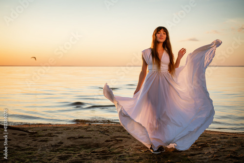 Beautiful young woman in a white long dress near the sea at sunset stands © Екатерина Переславце
