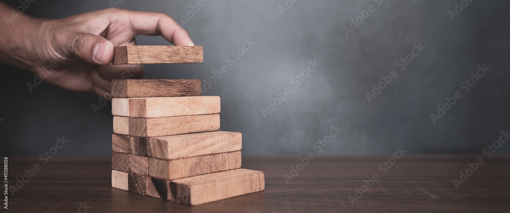 Close-up hand is placing wood block tower stacked in pyramid shape with caution to prevent collapse or crash concepts of financial risk management and strategic planning. Close-up hand is placing wood block tower stacked in pyramid shape with caution to prevent collapse or crash concepts of financial risk management and strategic planning.