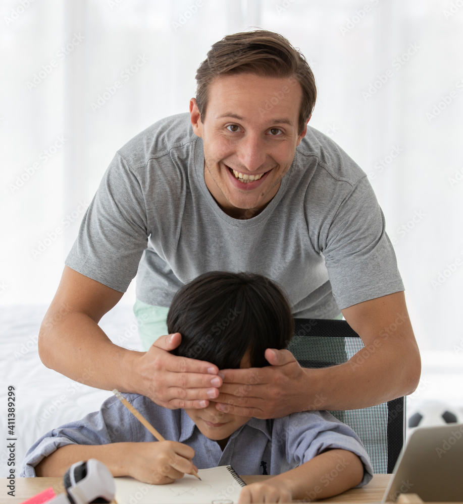Fototapeta premium Father with smile and fun face using hand to close eyes of mixed race son who sitting on desk