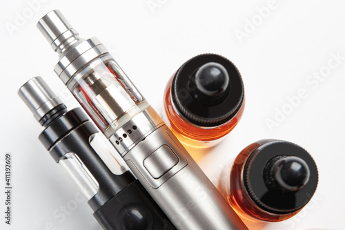 Silver and black vaping devices are nearby. Two e-cig next to vape oils. Vapor and smoking oils. Concept of replacing cigarettes with vaping devices. Smoking modern cigarettes. Modern e-cig close up