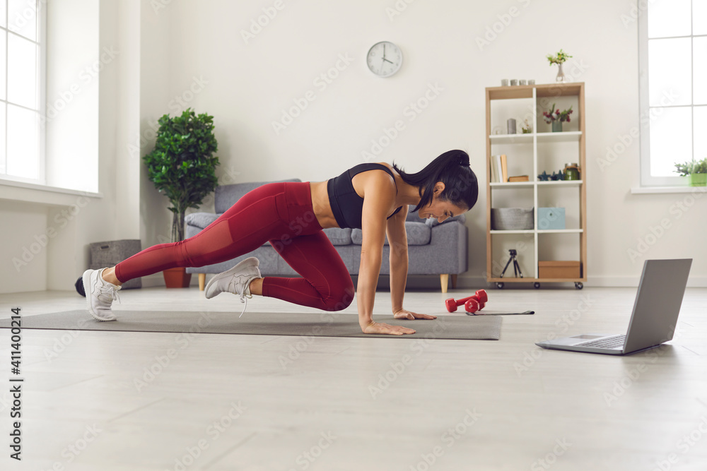 Side view of athletic woman in sports uniform and sneakers training on a mat in front of a laptop at home in the living room. Woman doing fitness exercises for legs using online training programs. Side view of athletic woman in sports uniform and sneakers training on a mat in front of a laptop at home in the living room. Woman doing fitness exercises for legs using online training programs.