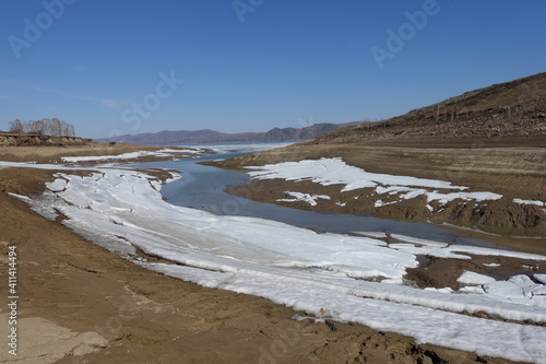 Harsh nature landscape and road through fields and mountains along the riverbed for off-road SUV with puddles and mud. Spring background with wet earth and snow and slush.