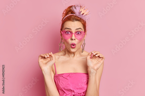 Surprised redhead young European woman wears trendy sunglasses raises hands stares at camera and says omg dressed in festive dress isolated over pink background. People style fashion concept © wayhome.studio 