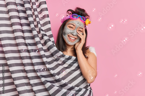 Positive Asian woman has smooth perfect skin applies clay mask smiles broadly applies hair rollers for making curly hairstyle poses behind curtain enjoys hygienic procedures poses around bubbles