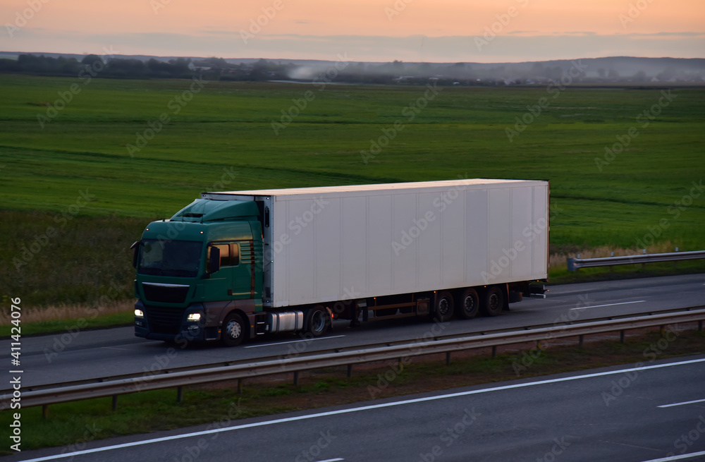 Truck with semi-trailer driving along highway on the sunset background. Goods delivery by roads. Services and Transport logistics. Soft focus. Object in motion.