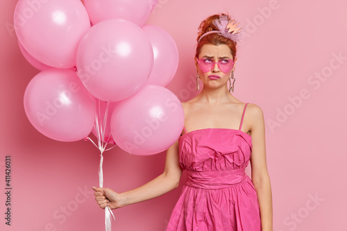 Displeased redhead woman dressed in nineties style outfit wears heart shaped sunglasses holds inflated balloons going to celebrate festive occasion isolated over pink background. Partying concept