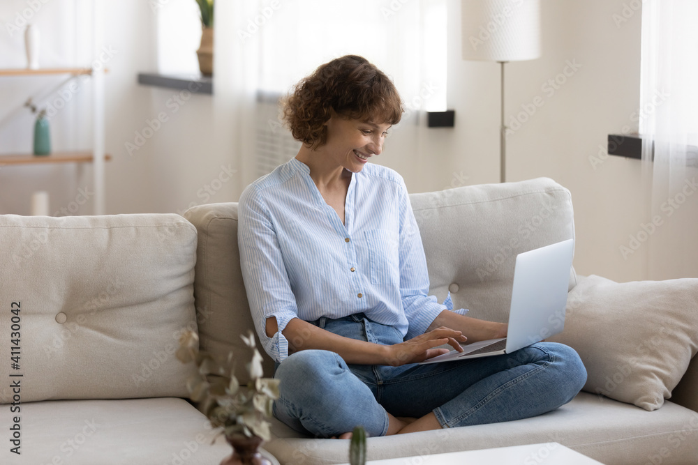 Smiling young Caucasian woman sit relax on couch in living room work online on laptop gadget from home office. Happy female rest on sofa browse wireless internet, text or message on web. Smiling young Caucasian woman sit relax on couch in living room work online on laptop gadget from home office. Happy female rest on sofa browse wireless internet, text or message on web.