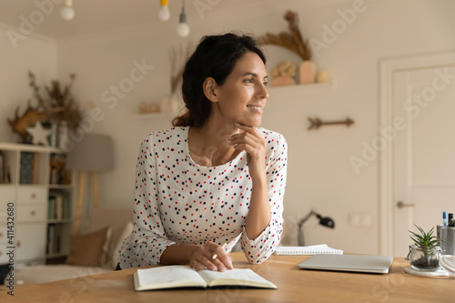Photos Smiling young Caucasian woman sit at table at home write make list in notebook look in distance dreaming thinking
