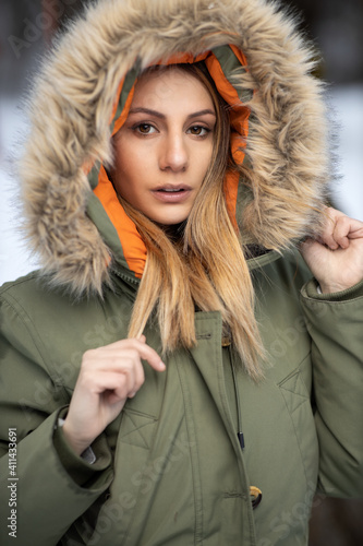 portrait of blonde girl in a green winter coat with a hood with furry fur on the snow in the mountains in winter