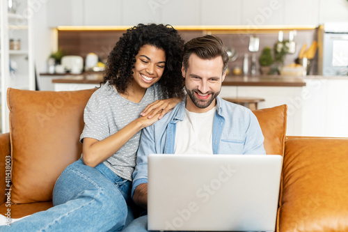 Multi ethnic couple in love spends leisure time with a laptop at home. Multiracial girlfriend and caucasian boyfriend look at the laptop screen sitting on the sofa, watching movies