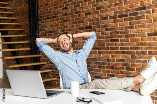 Cheerful young businessman leaned back in the chair, holds his hands behind his head and looks at laptop. Smiling freelancer feeling satisfaction after successful working day, good deal concept