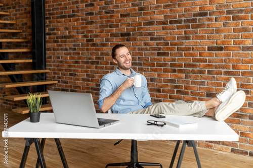 Successful male entrepreneur takes a break on the workplace, hipster guy sits in relaxed pose with his feet on the table and resting with a cup of coffee in the modern office space photo